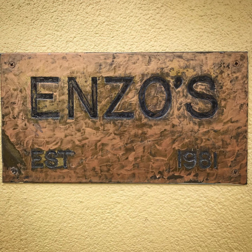 Weathered copper sign reading Enzo's, Est. 1981, mounted on a yellow stucco wall