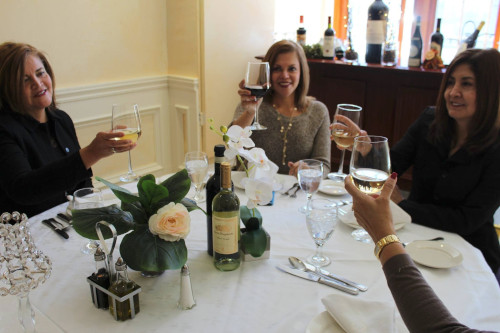 Three women toasting with wine glasses at a table with a rose centerpiece